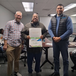 Three men standing together smiling. The one in the middle is holding a certificate that he just received