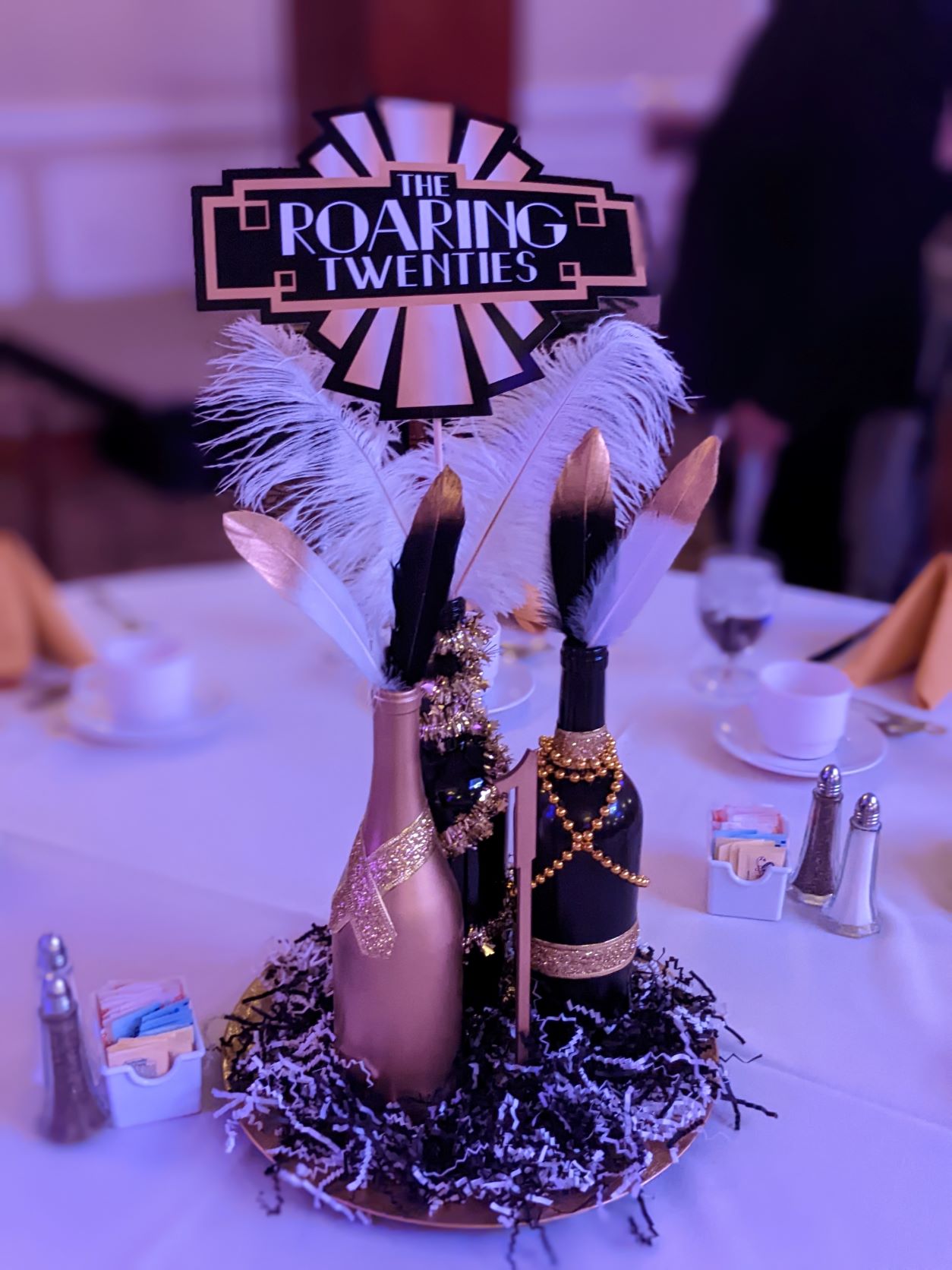 Centerpiece on round 10-top with three champagne bottles with beads and feathers, colors in black and gold