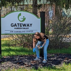 Woman bent over picking up litter in front of Gateway Hudson Valley sign